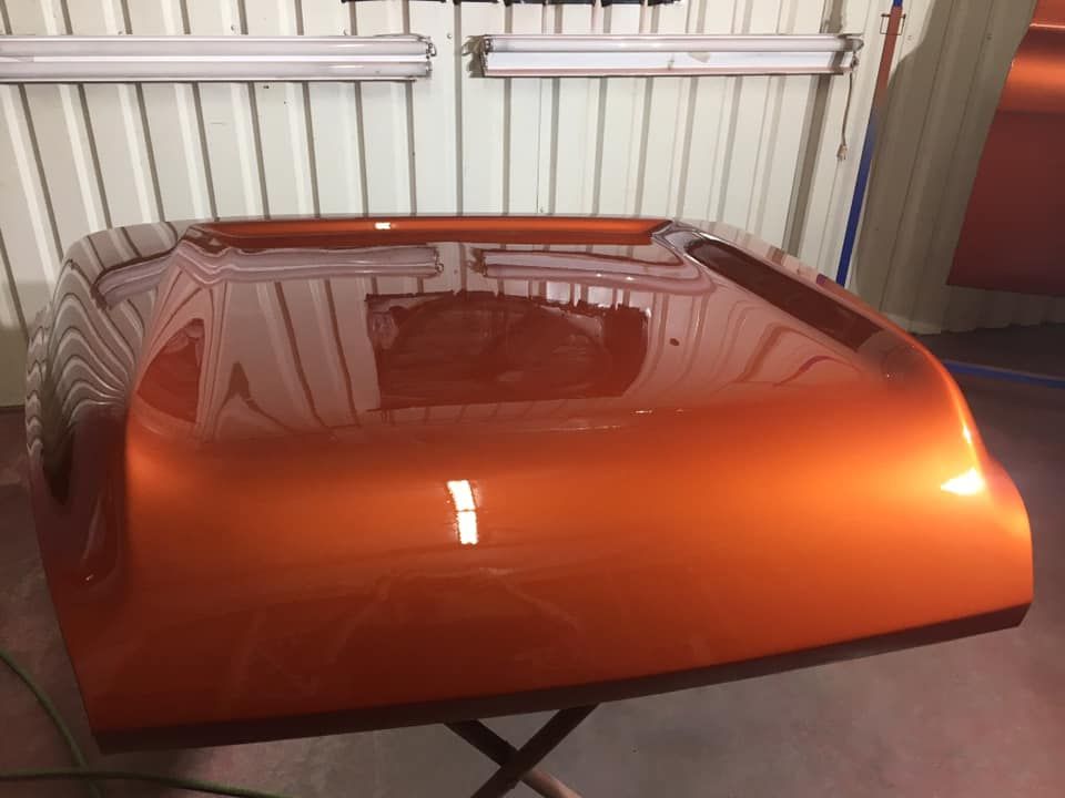 Orange car hood, freshly painted with a glossy finish, sits on a stand in a workshop.