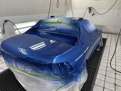 Blue car being painted in a paint booth, covered in plastic and masking tape.