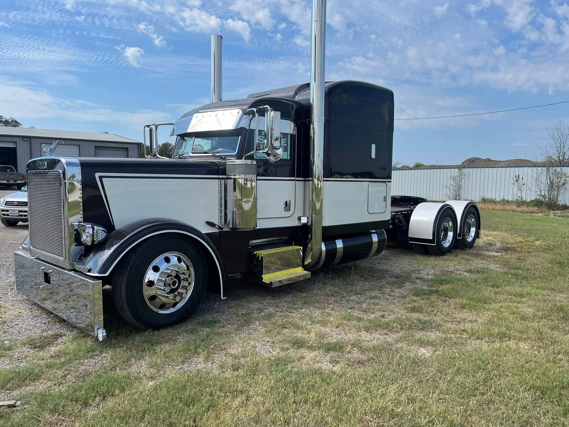 Black and white semi-truck parked outdoors. Silver grill and stacks, chrome wheels, blue sky background.