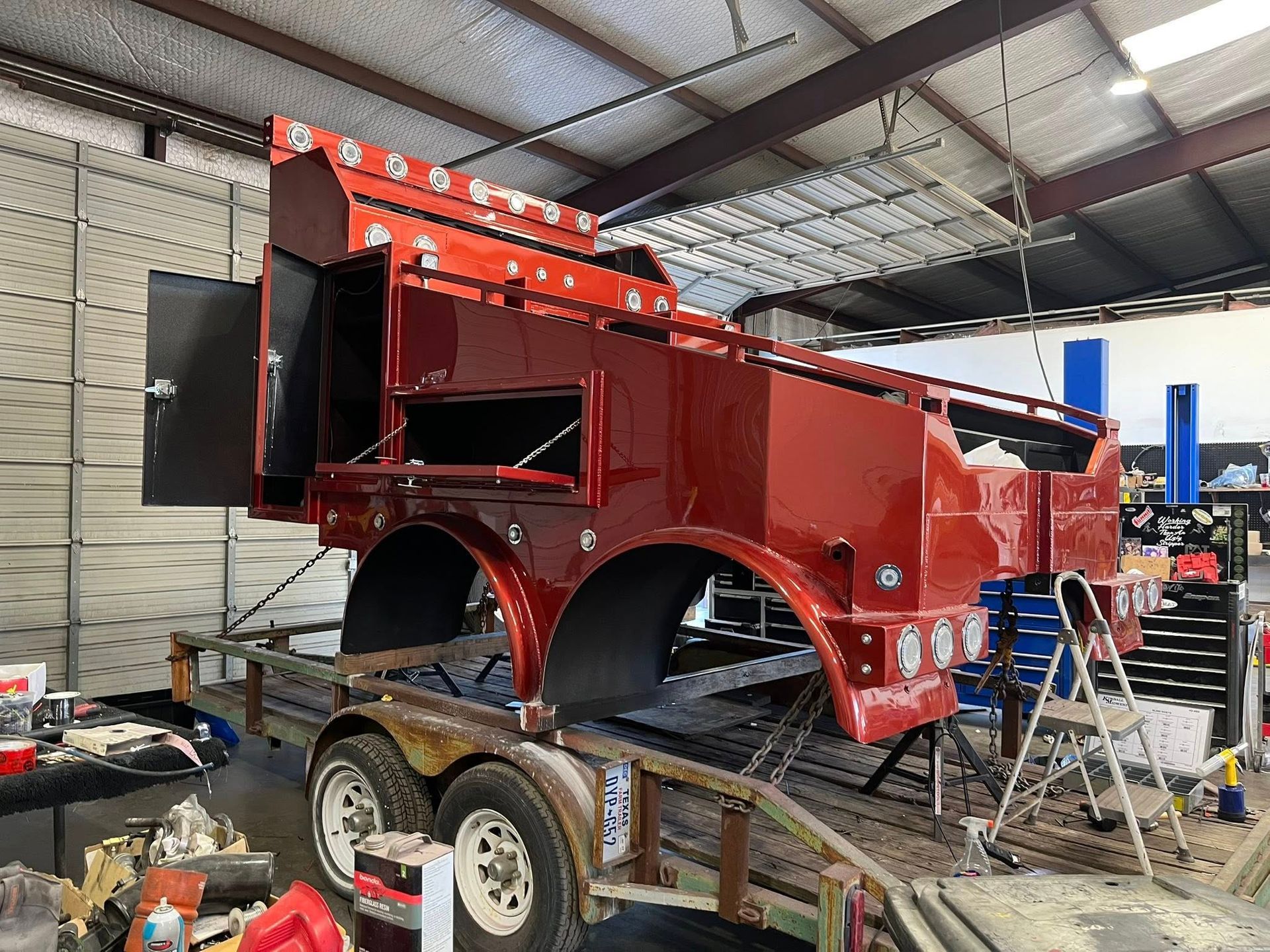 Red metal truck bed on a trailer inside a workshop. Open side compartments and wheel wells are visible.