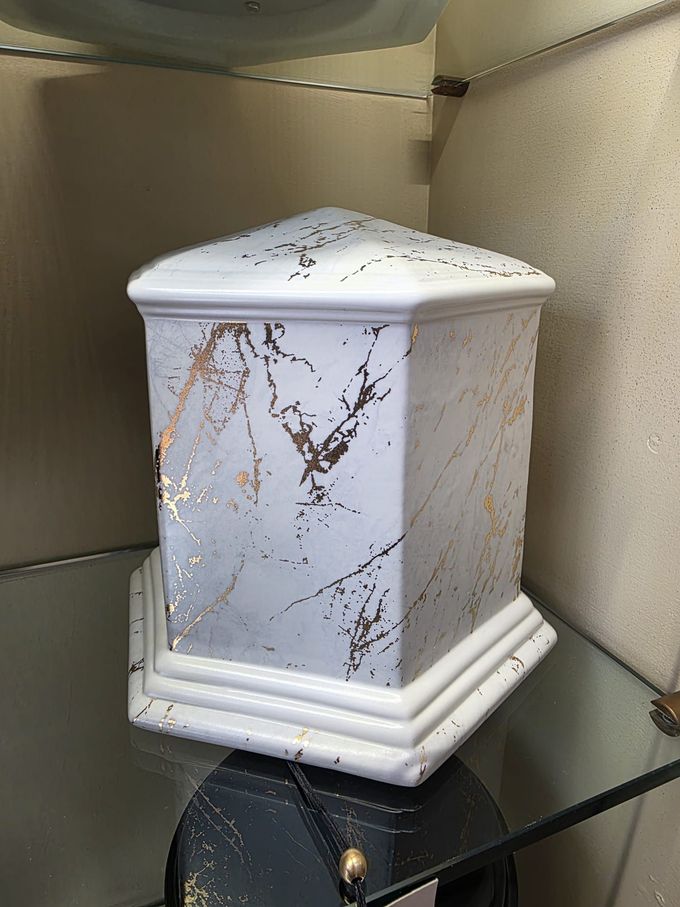Un'urna funeraria bianca con una fascia rossa è appoggiata su una superficie di legno accanto a una cornice e a una pila di libri.