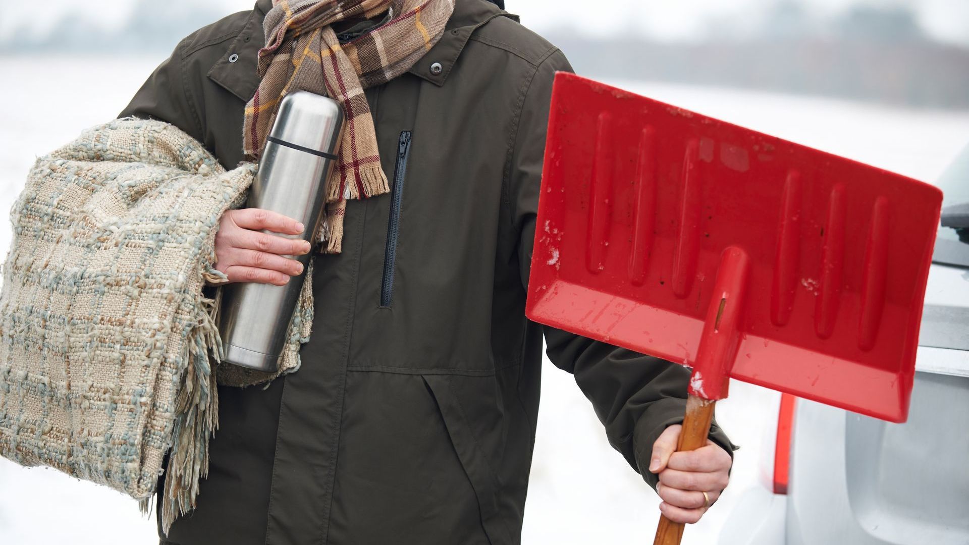 A person in a coat and scarf is holding a blanket, thermos and shovel. The weather is snowy and cold.