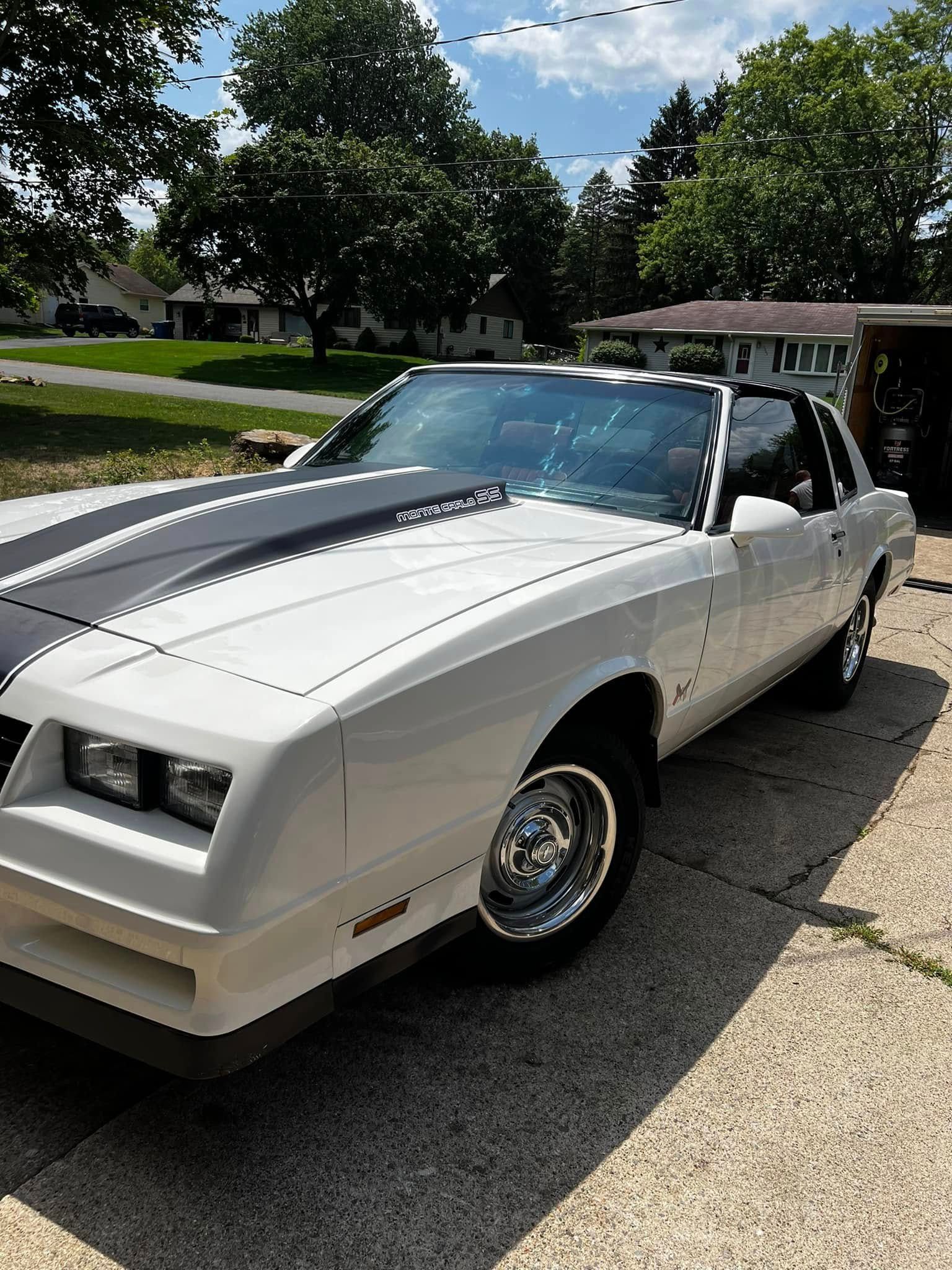 A white Chevrolet Monte Carlo with black racing stripes parked on a paved driveway on a sunny day.