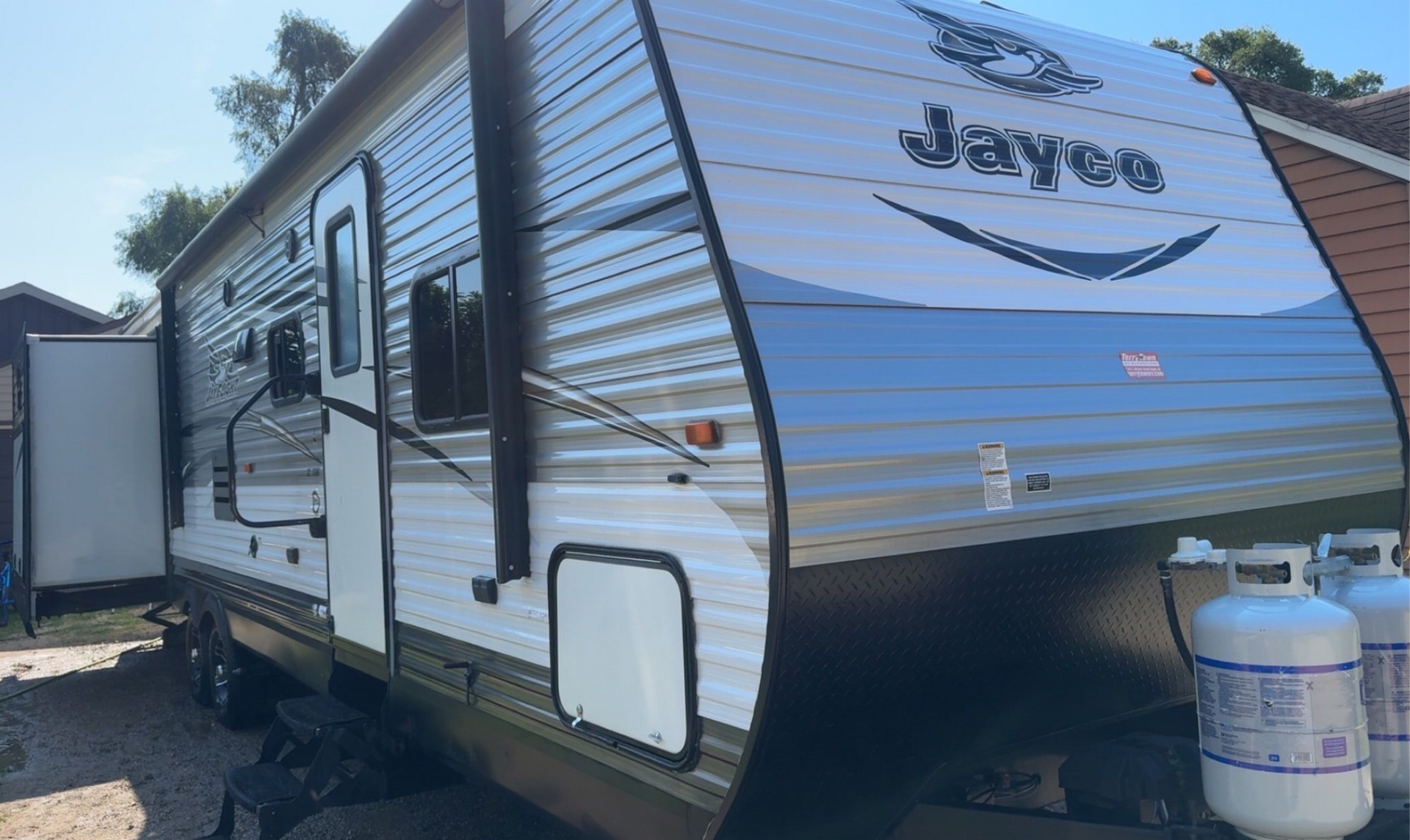 White and gray Jayco travel trailer parked outdoors beside a propane tank on a sunny day