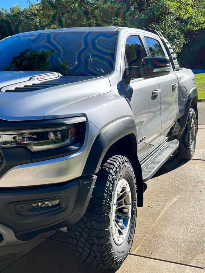 A silver Ram TRX pickup truck parked on a concrete driveway with large off-road tires and a black hood scoop.