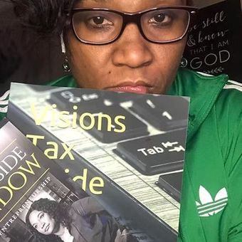 Person in glasses holding several books, with a chalkboard in the background.