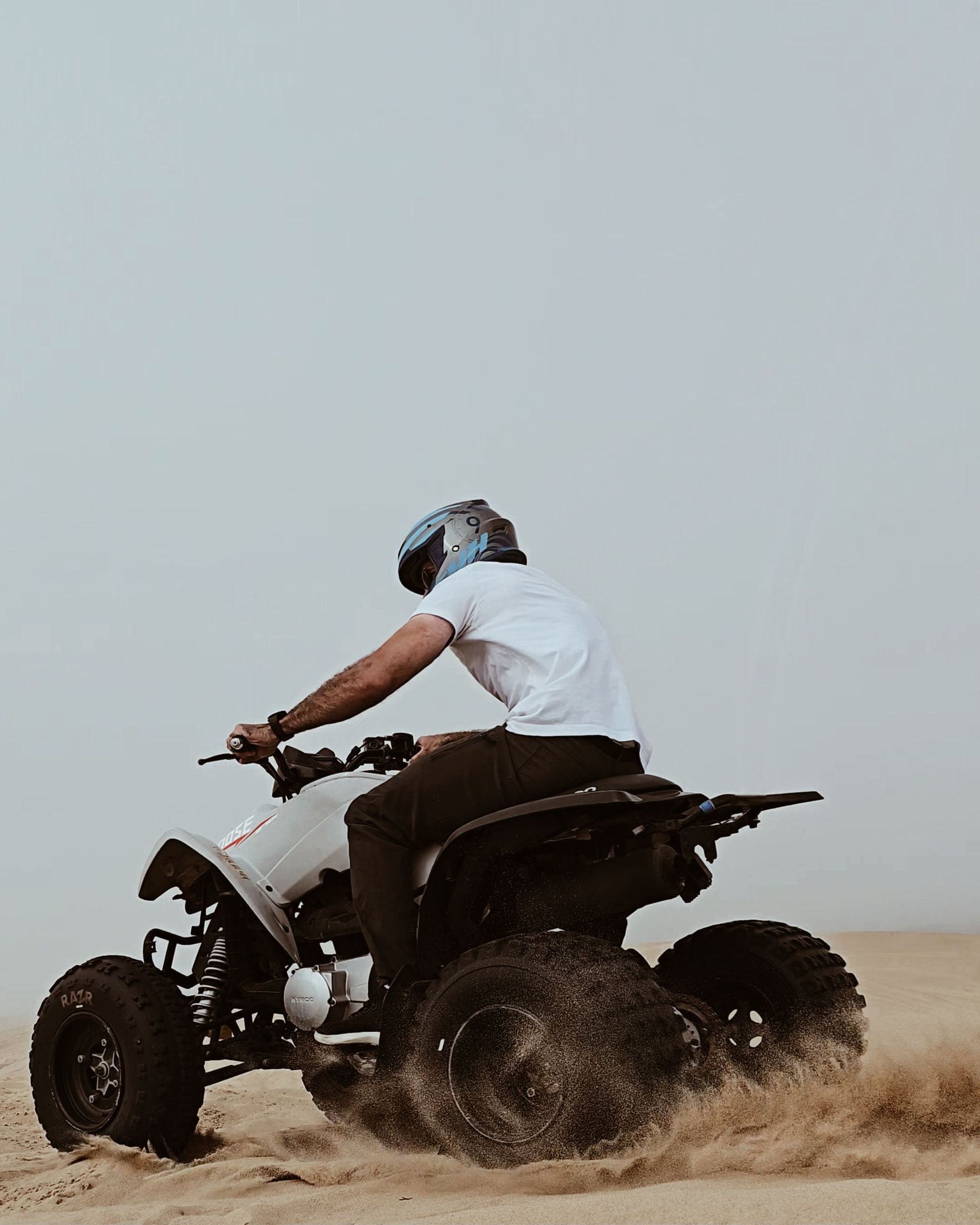 Person riding an ATV through sand, kicking up dust under a gray sky