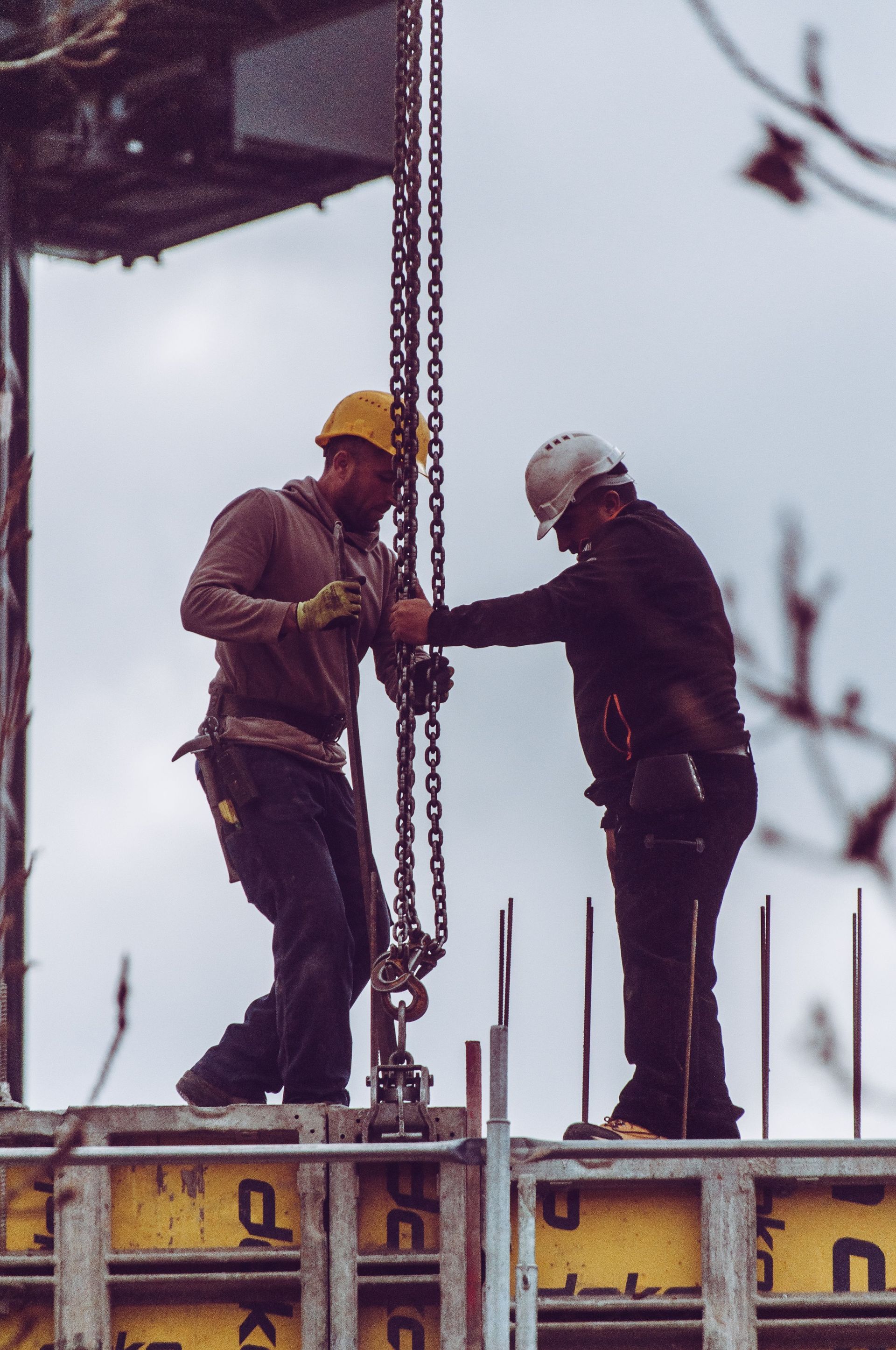 Two construction workers on scaffolding beside a vertical beam, wearing hard hats and safety gear.