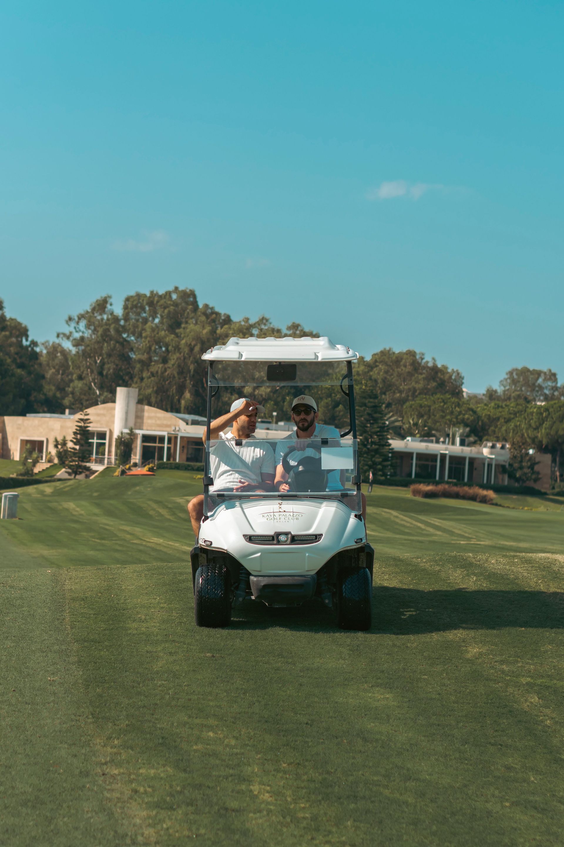 Two people driving a golf cart on a grassy course under a clear blue sky