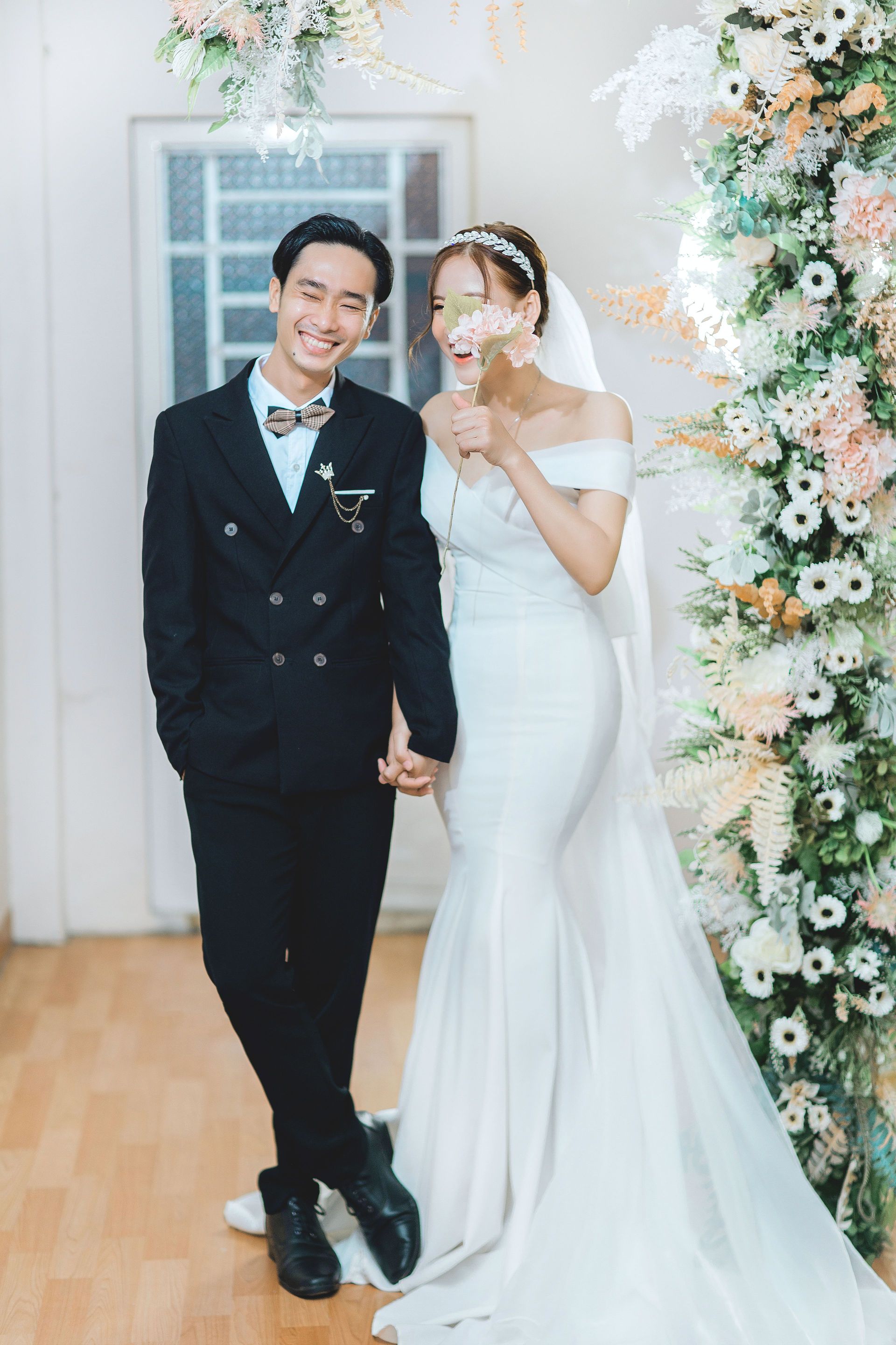 Bride and groom smiling in wedding attire, standing under a floral arch indoors