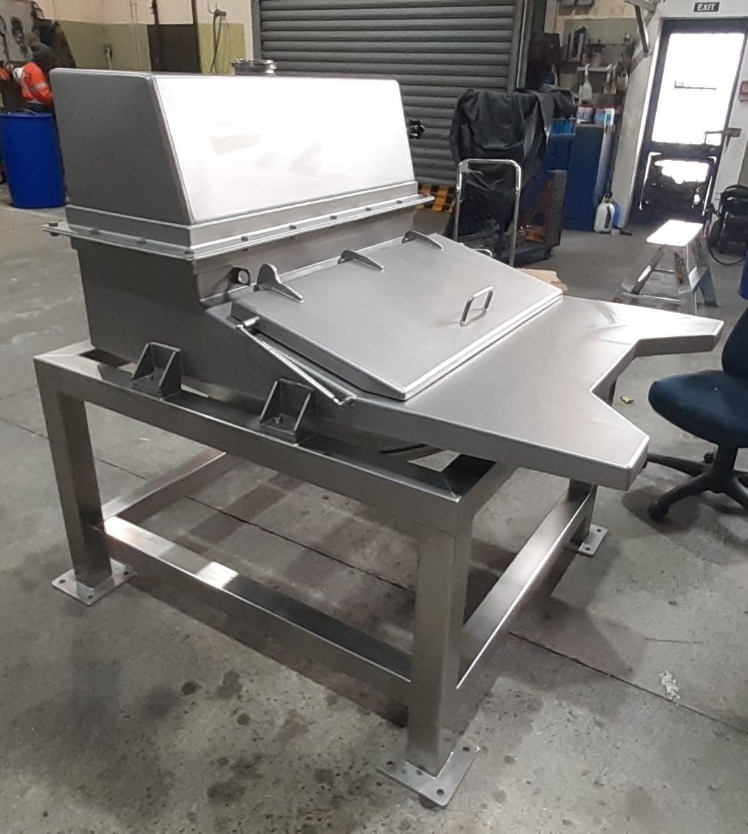 Stainless steel industrial machine with a tilting tray on a metal frame, inside a workshop.
