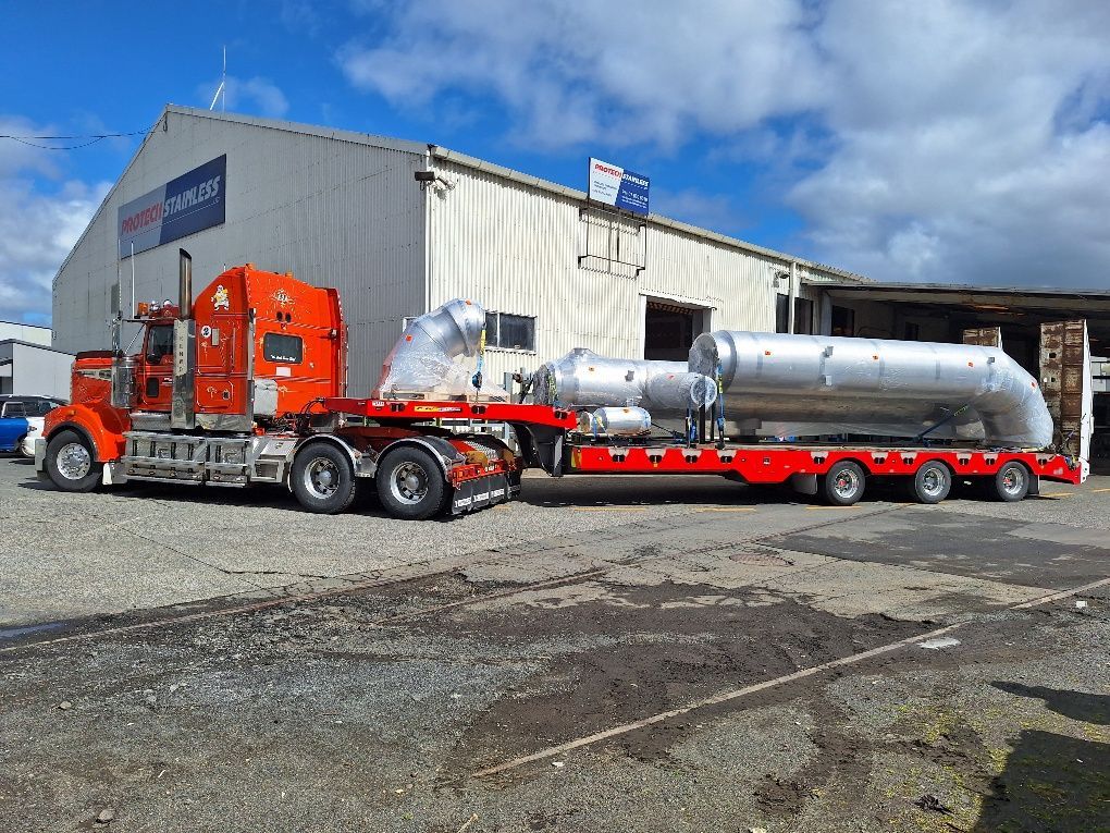 An orange semi-truck pulling a flatbed trailer with large, silver industrial equipment outside a warehouse. An orange semi-truck pulling a flatbed trailer with large, silver industrial equipment outside a warehouse.