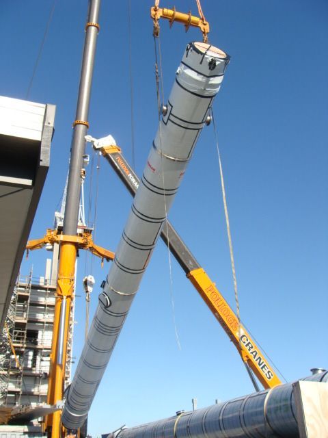 Two cranes lift a long, cylindrical white industrial component against a blue sky.