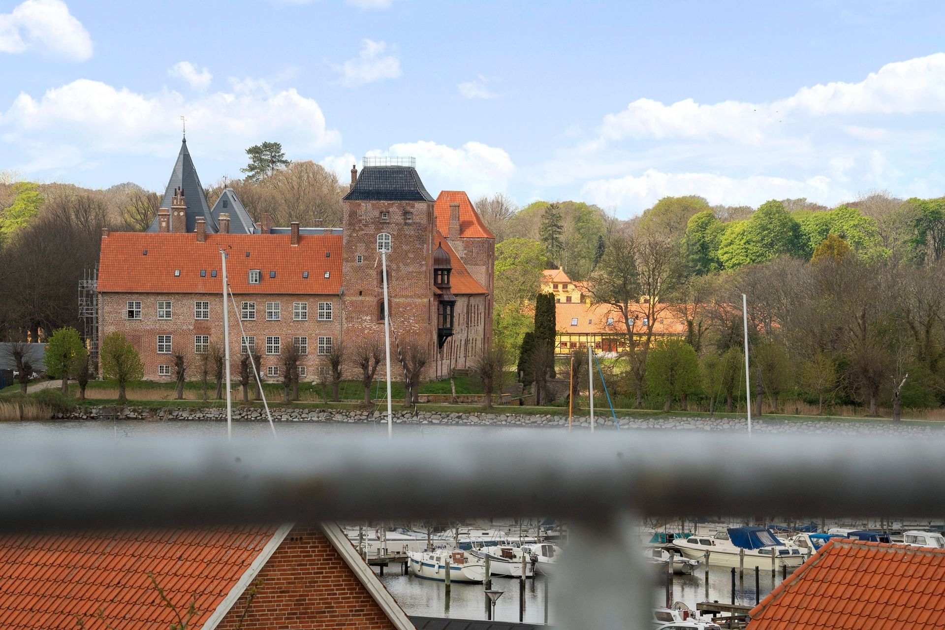 Slot med røde tegltage, brune stenmure og et højt tårn, mod en baggrund af træer og en blå himmel.