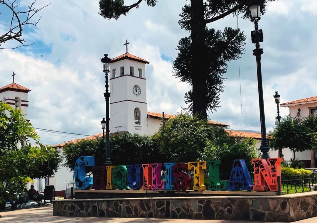 Letrero colorido de “TUXTLA” en una plaza con una torre de reloj blanca y árboles bajo un cielo azul.