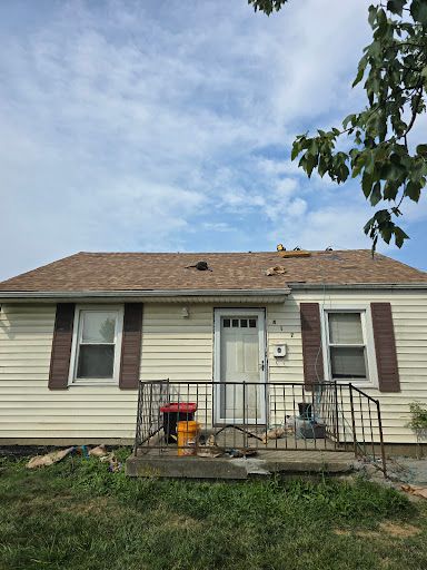 Small beige house with brown shutters, front porch railing, and a cluttered roof under a cloudy sky.