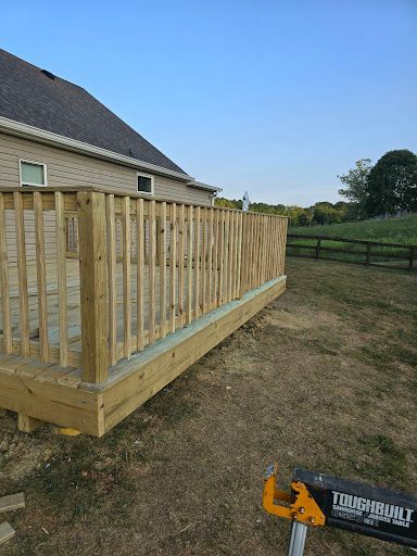 Wooden deck railing under construction beside a house, with a drill on the ground in a grassy yard.