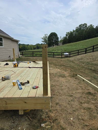 Wooden deck under construction beside a house, with tools scattered and a fenced grassy slope beyond.