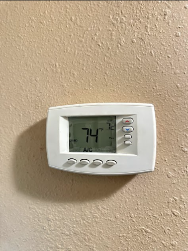 White thermostat mounted on a textured tan wall, displaying 74 degrees with AC activated.