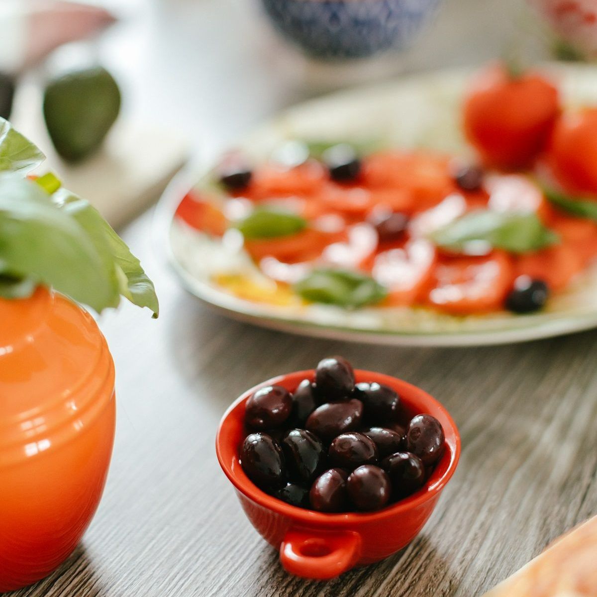 A bowl of olives sits on a table with a plate of food in the background