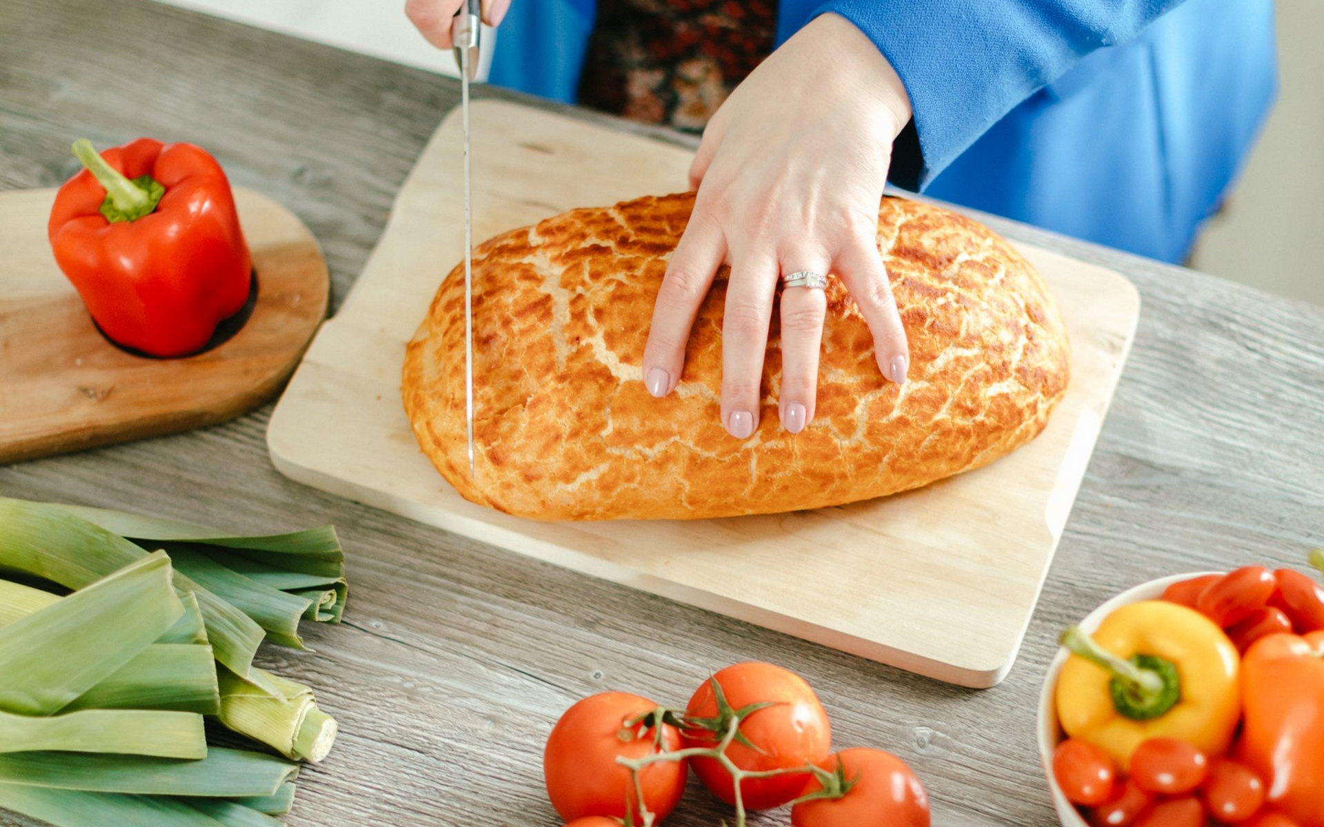 Person cutting bread on a wooden board with various vegetables nearby.