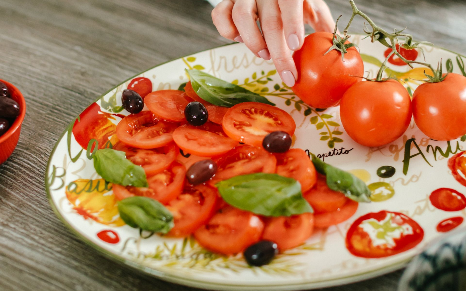 Tomato and basil salad with olives on a decorative plate, person adds tomatoes.