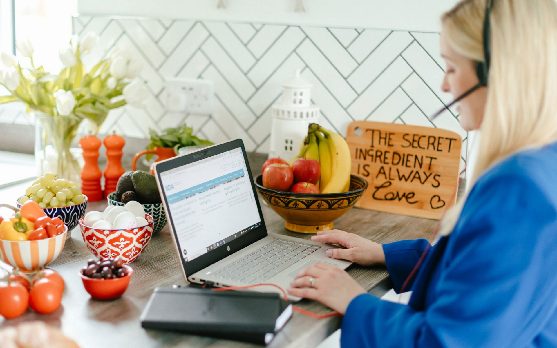 Woman with headset working on laptop in kitchen, surrounded by fruits, vegetables and a sign.