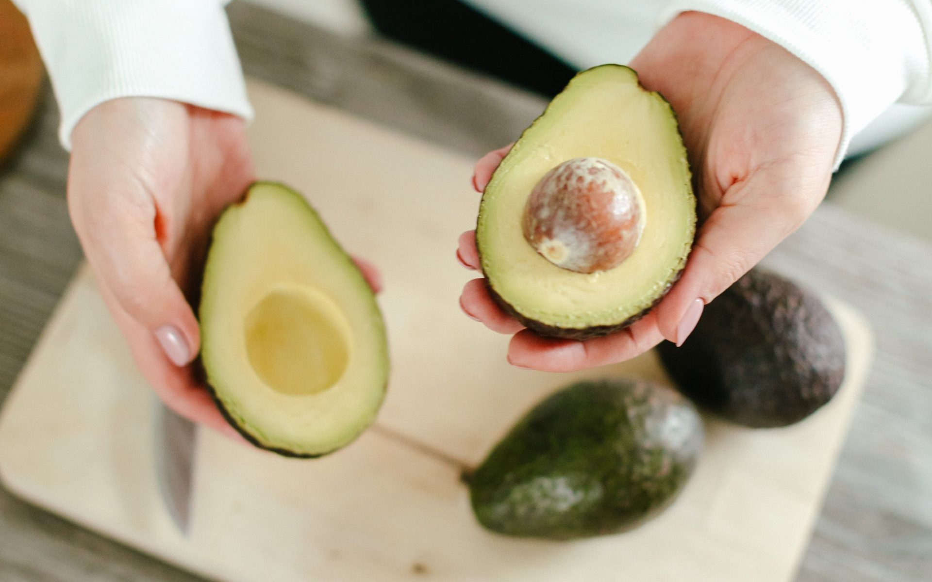 Hands holding two avocado halves, one with the pit. Two whole avocados and a knife on a cutting board.