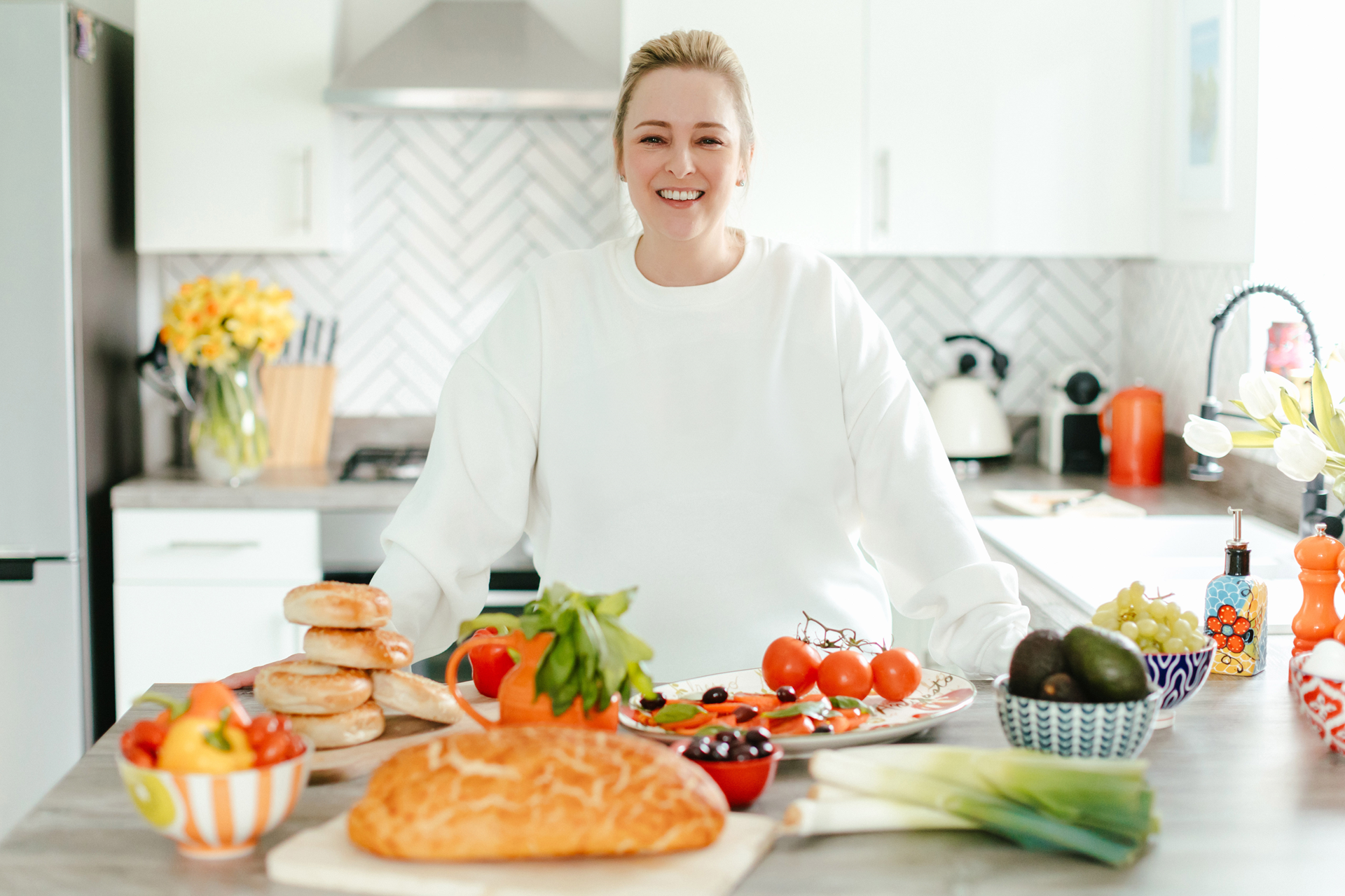 Woman smiles in kitchen, surrounded by food: bread, vegetables, and fruit.