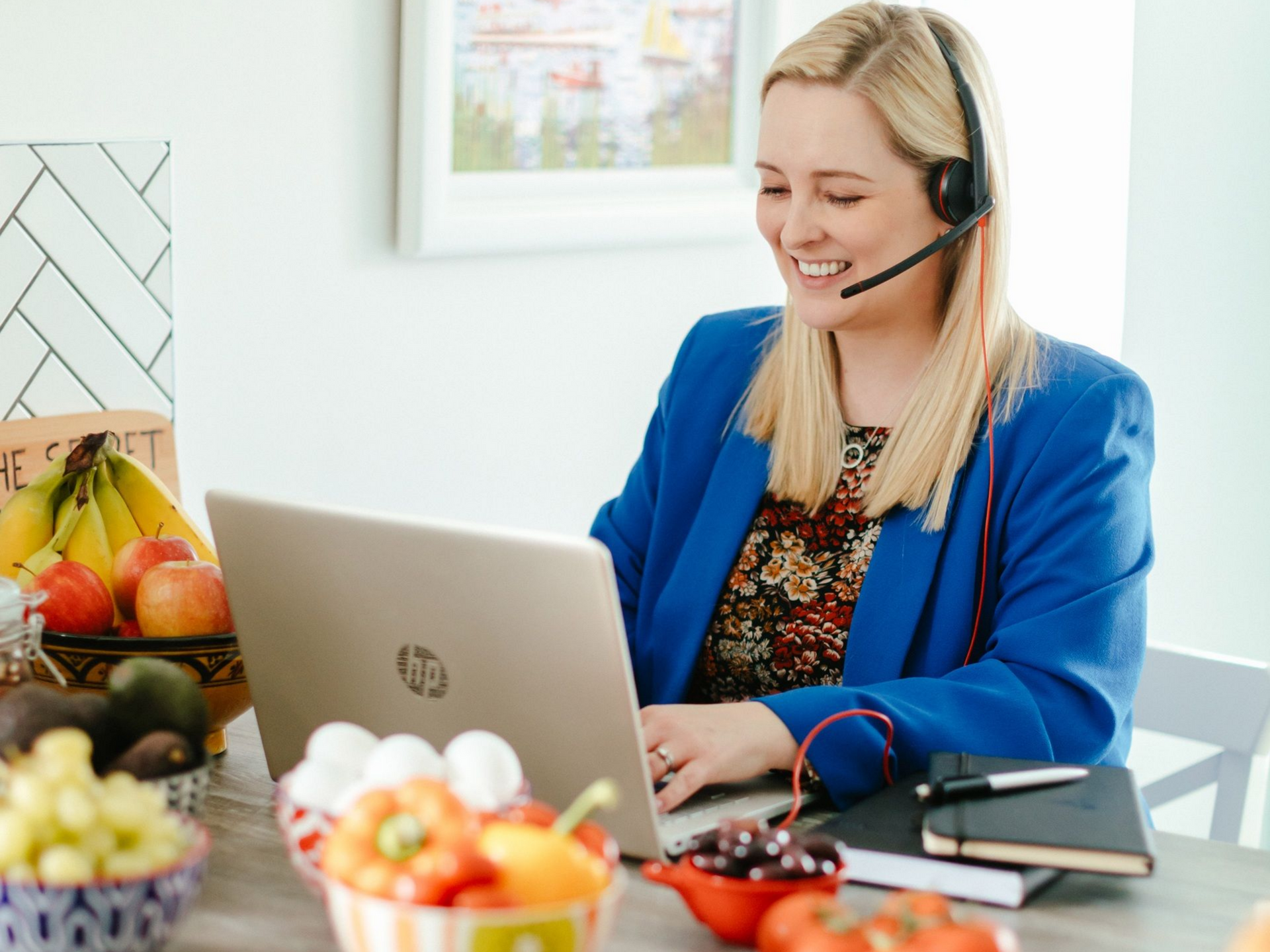 Woman with headset smiles while working on laptop at kitchen table with food.