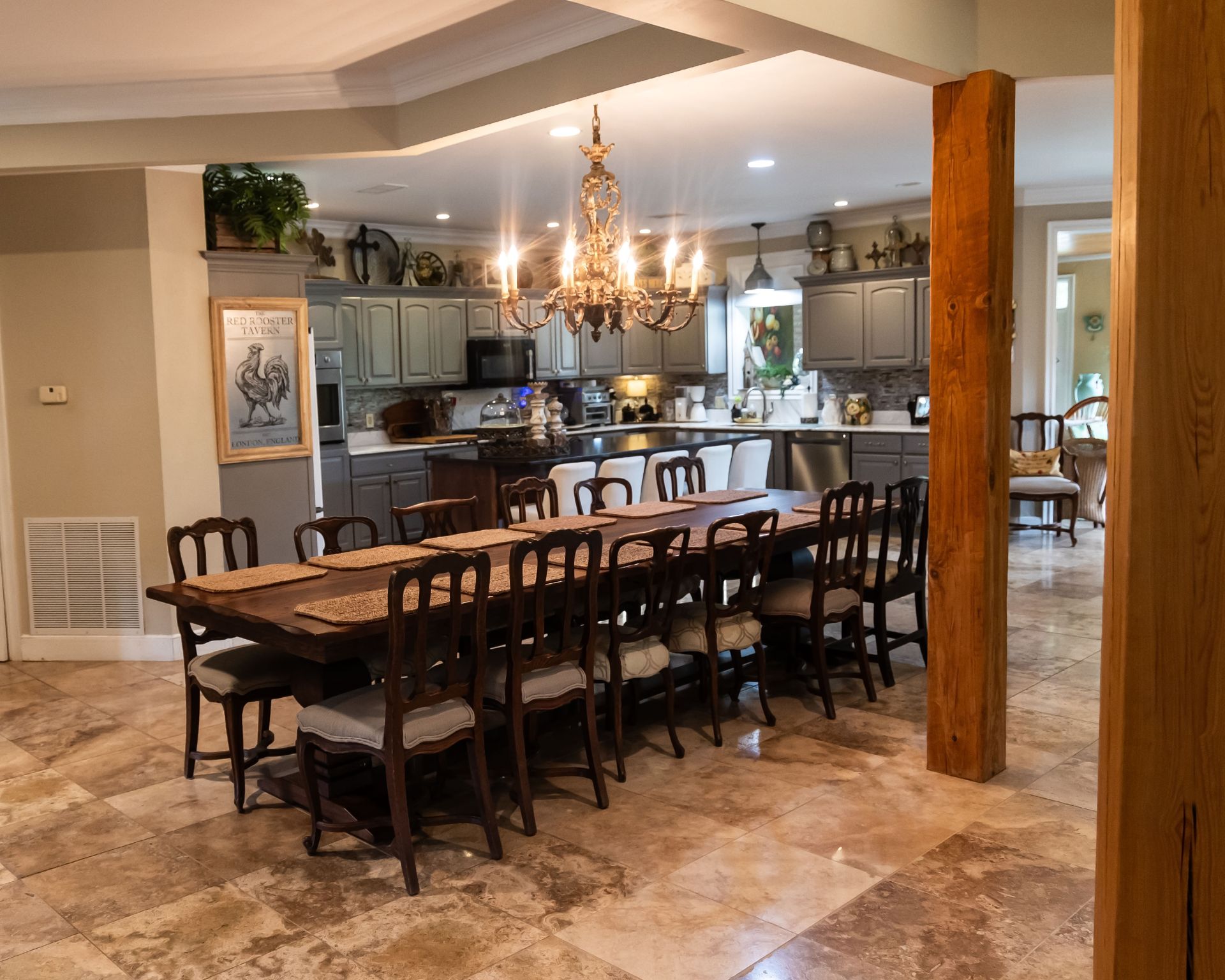 Elegant dining room with long wooden table, chandelier, tiled floor, and open view into a kitchen.