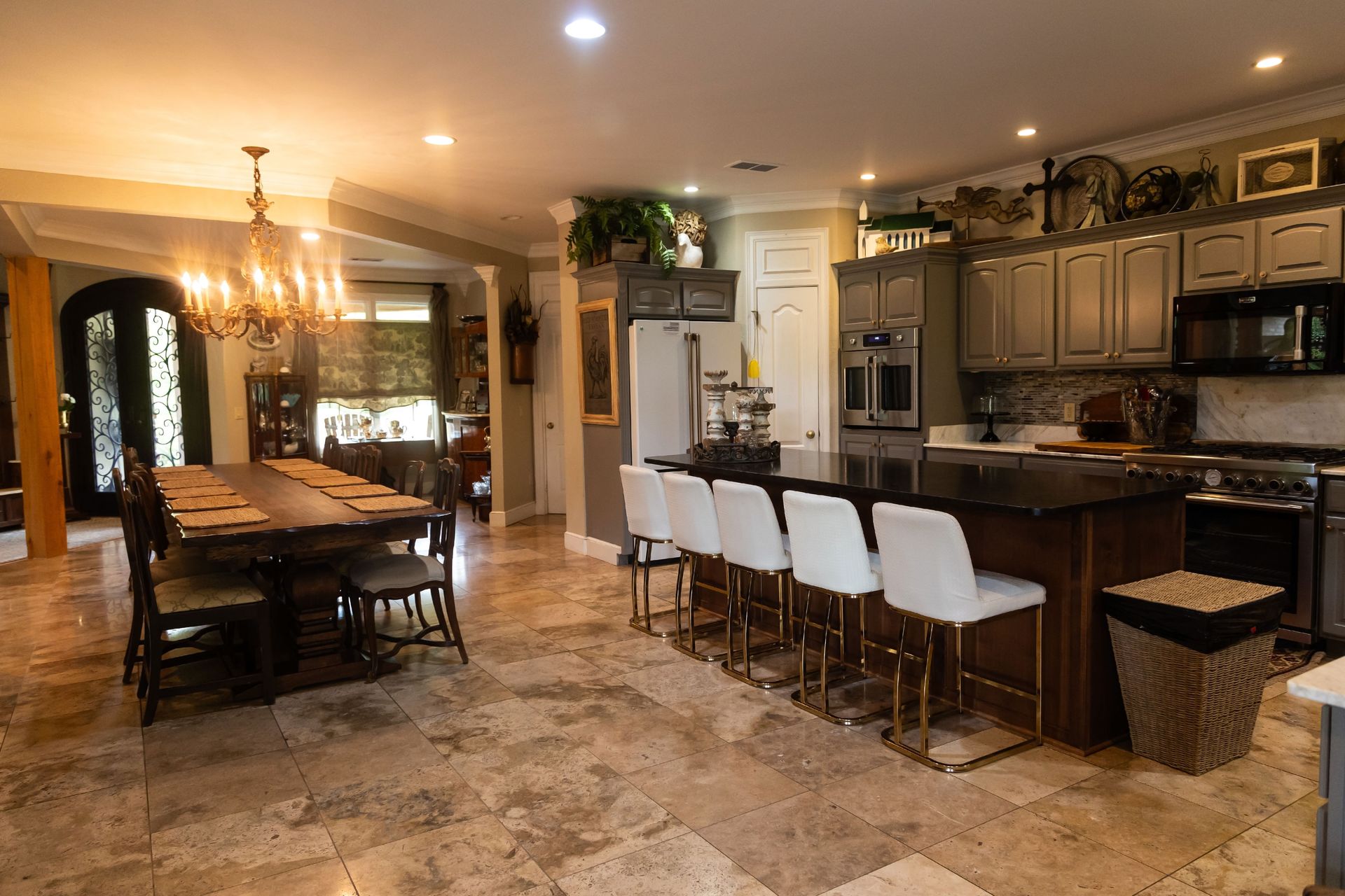 Spacious kitchen with dark island, white stools, marble floors, and a dining table under warm lighting