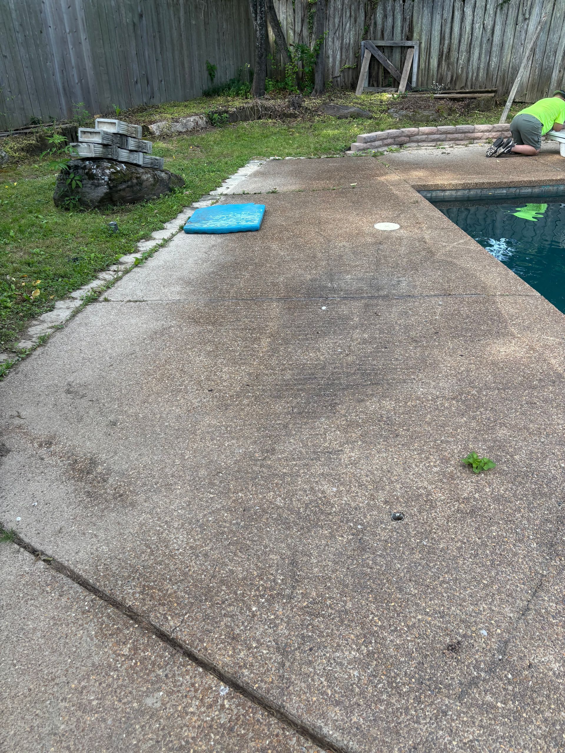 Backyard concrete patio beside a pool, with a blue cushion and a person in a green shirt at the edge