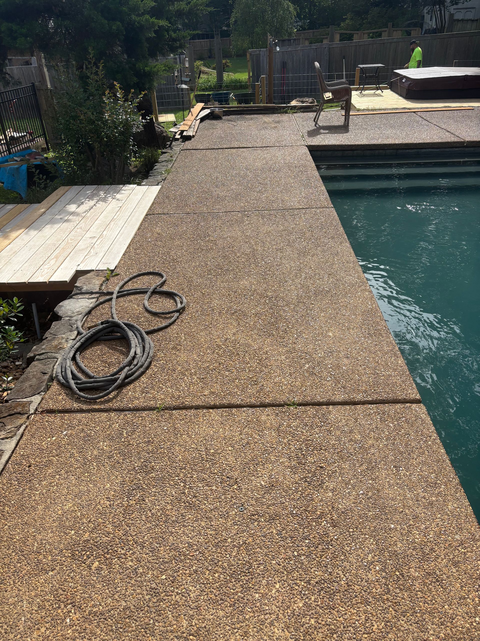 Poolside pebble walkway beside blue water, with coiled hose and wooden bench in a sunny backyard
