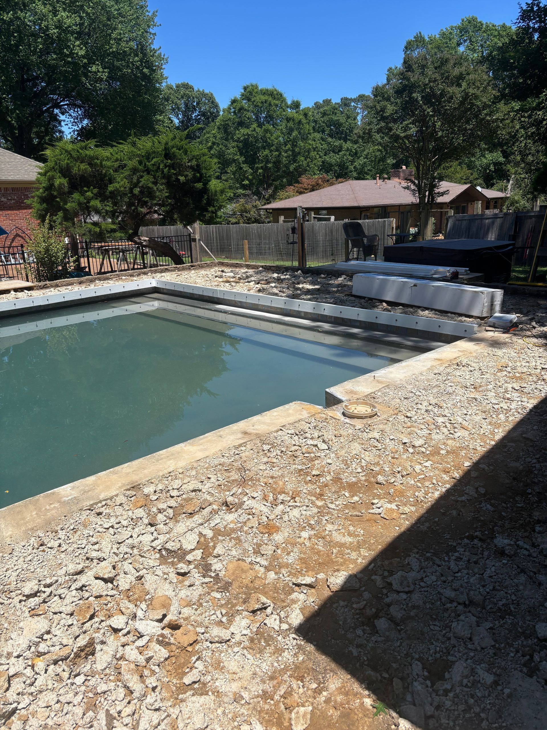 Empty backyard pool with cloudy water, stone deck, and shaded seating under trees on a sunny day