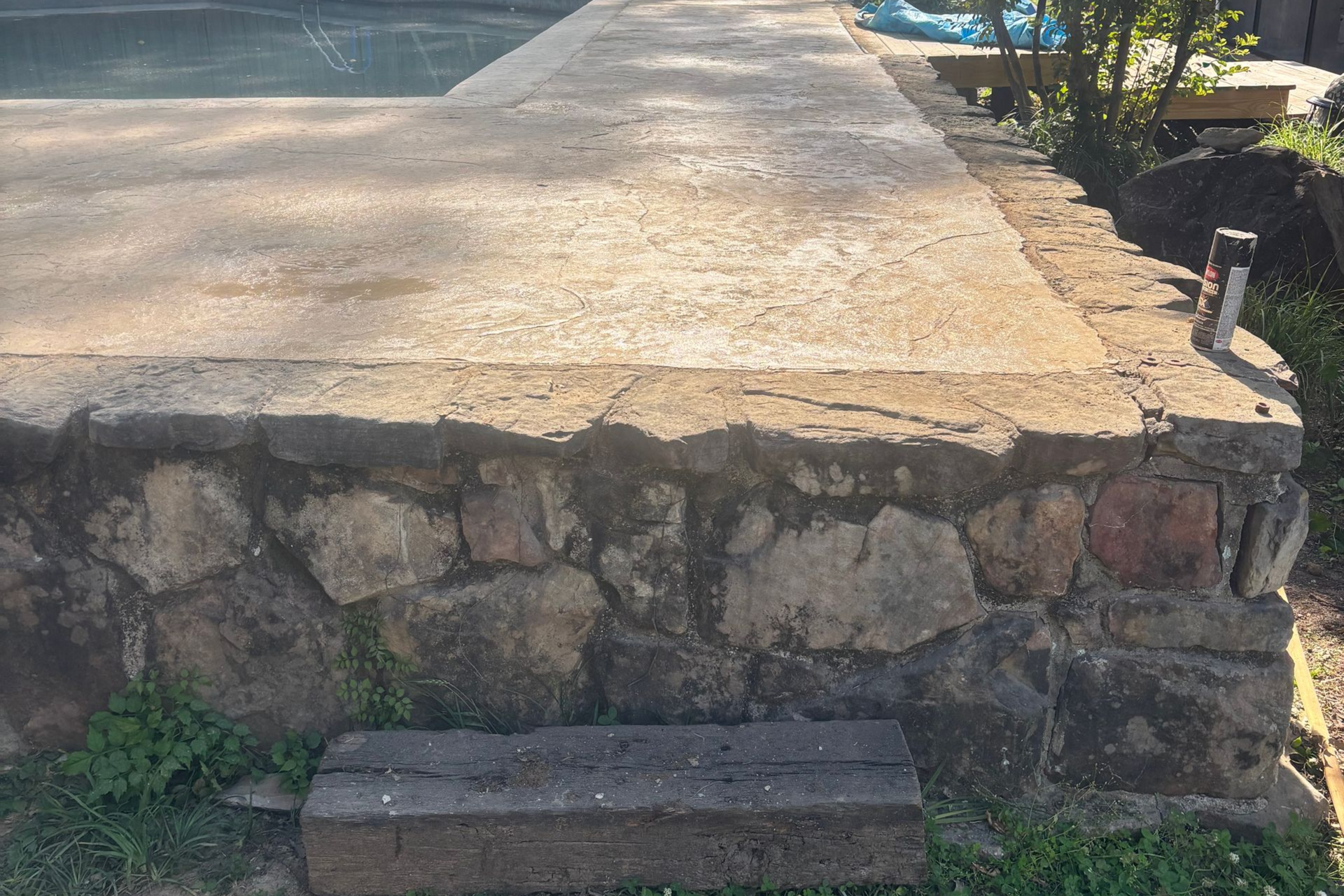Stone retaining wall supporting a cracked concrete path beside a grassy area and shrubs