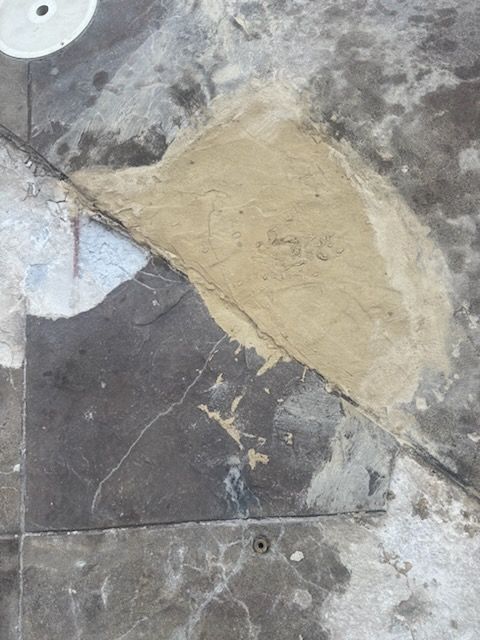 Rough concrete surface with a tan patch repair over dark gray and white stone tiles.