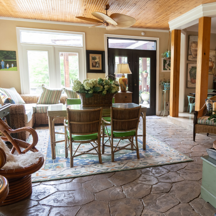 Cozy living room with wicker chairs, dining table, stone floor, and large windows letting in daylight