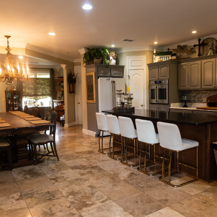 Warm kitchen with dark wood island, white bar stools, tiled floor, and adjacent dining area under recessed lighting