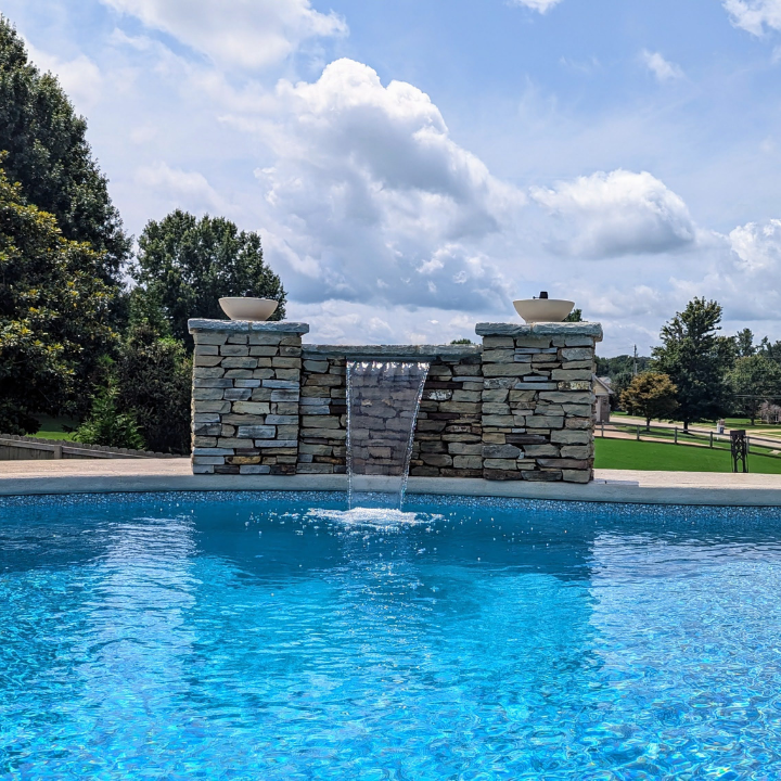 Blue swimming pool with a stone waterfall fountain under a cloudy sky