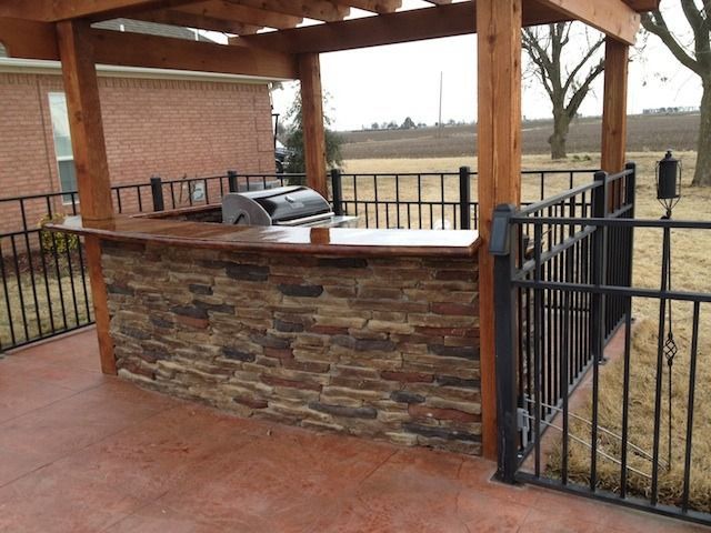 Covered outdoor grilling area with stone counter, built-in grill, and black metal railing overlooking a yard