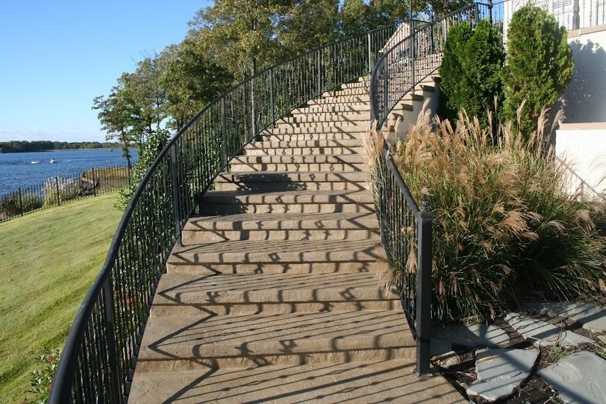 Curved wooden stairs beside a waterfront path with black railings and plants under a clear sky