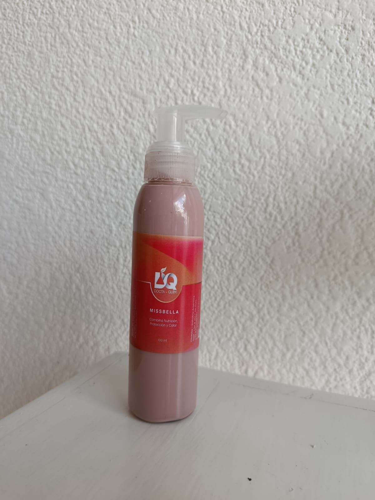 Botella de loción rosa con dispensador de bomba, sobre una superficie blanca contra una pared texturizada.