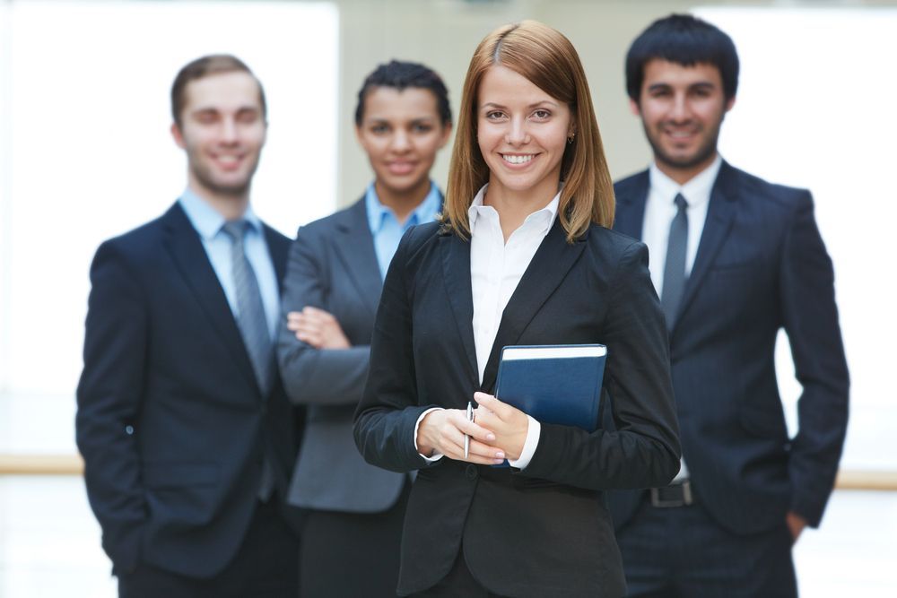 Cuatro profesionales de negocios con traje, de pie juntos; una mujer sostiene una carpeta y sonríe a la cámara