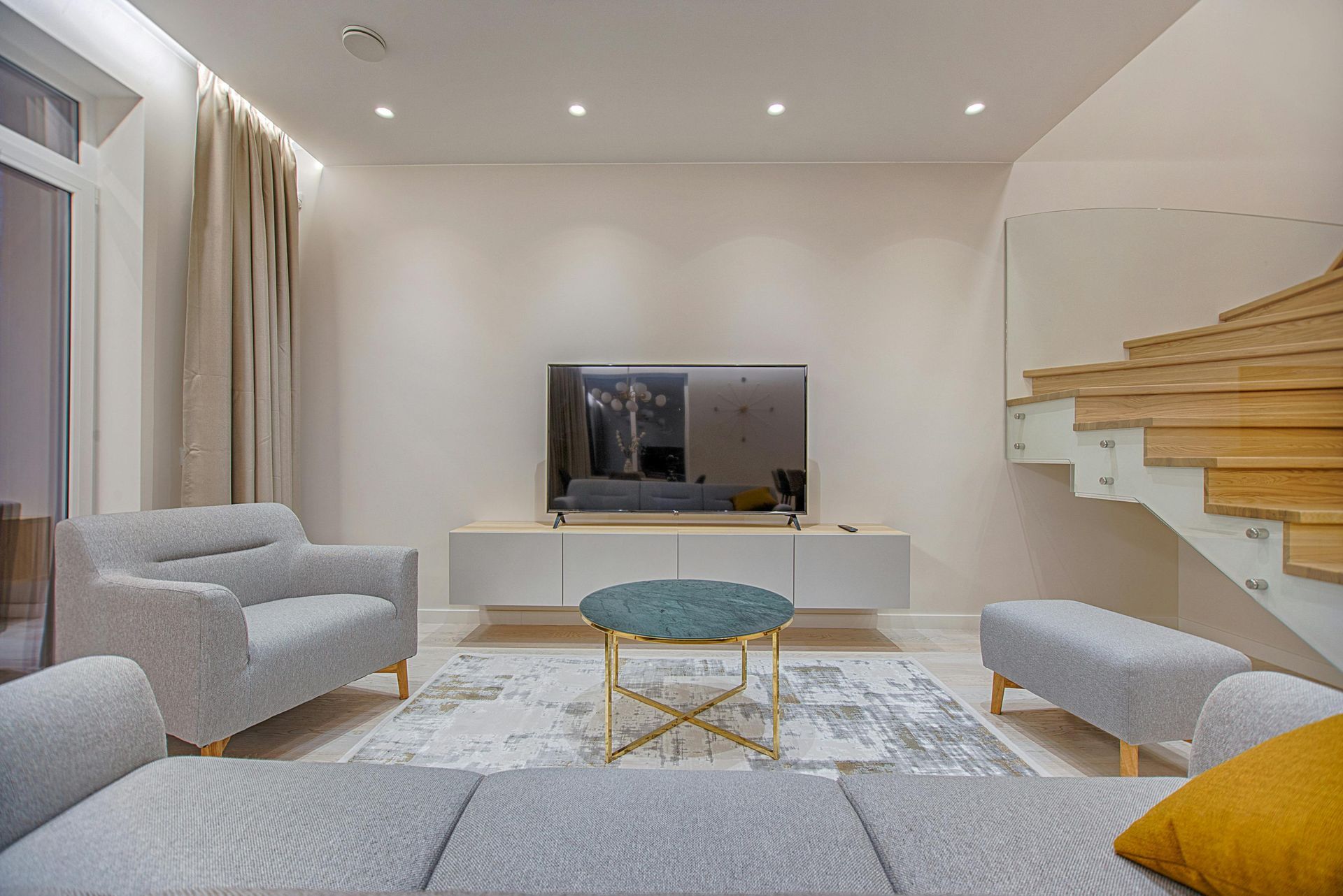 Modern living room with gray seating, TV on a white console, round coffee table, and staircase on the right