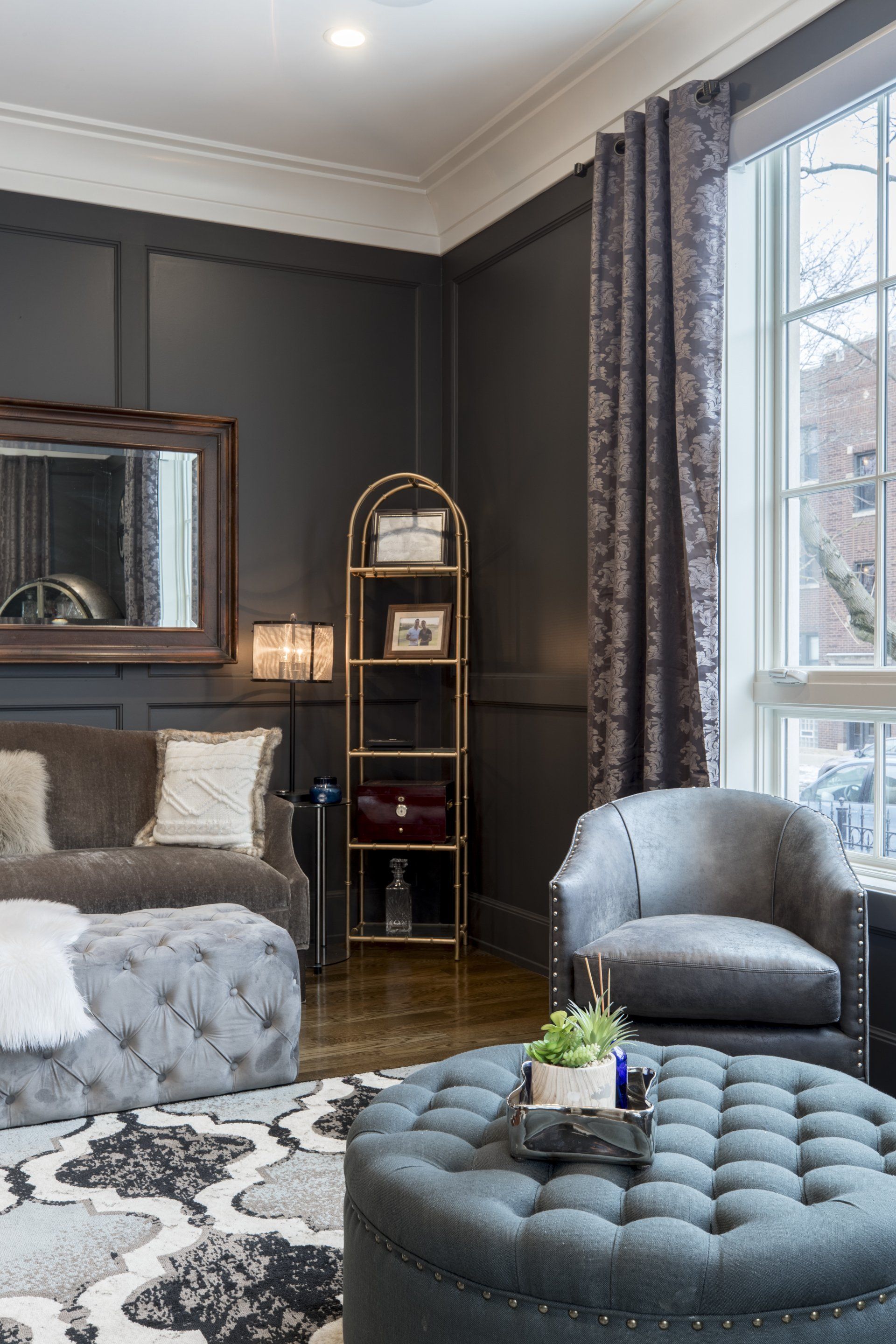Elegant bedroom with gray walls, tufted ottoman, armchair, bed, mirror, and tall window with patterned curtains