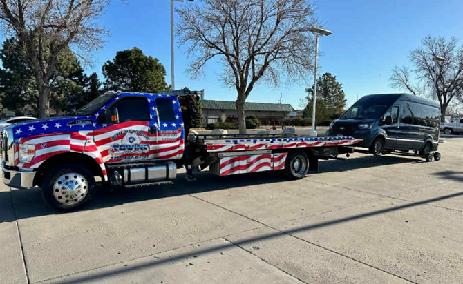 A tow truck with an American flag design hauling a dark-colored van. Outdoors, daytime.