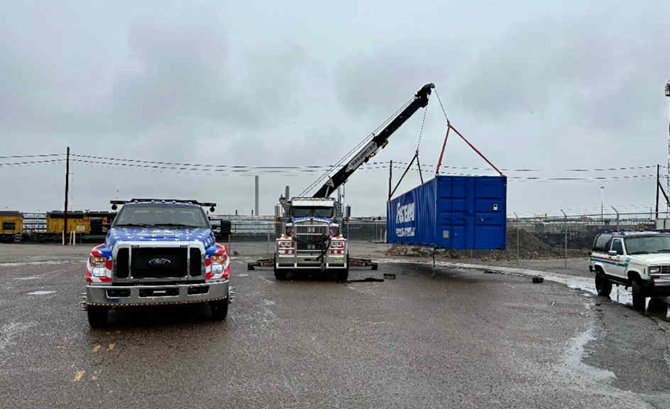 A tow truck crane lifting a blue shipping container in an industrial setting, overcast day.