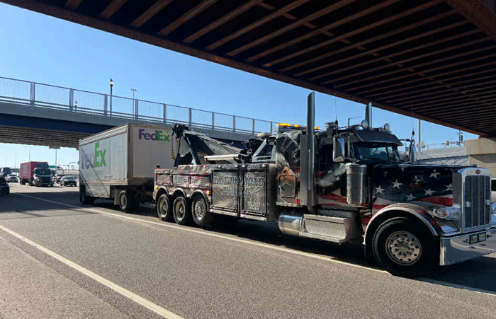 Tow truck with a FedEx trailer under a bridge on a sunny day.