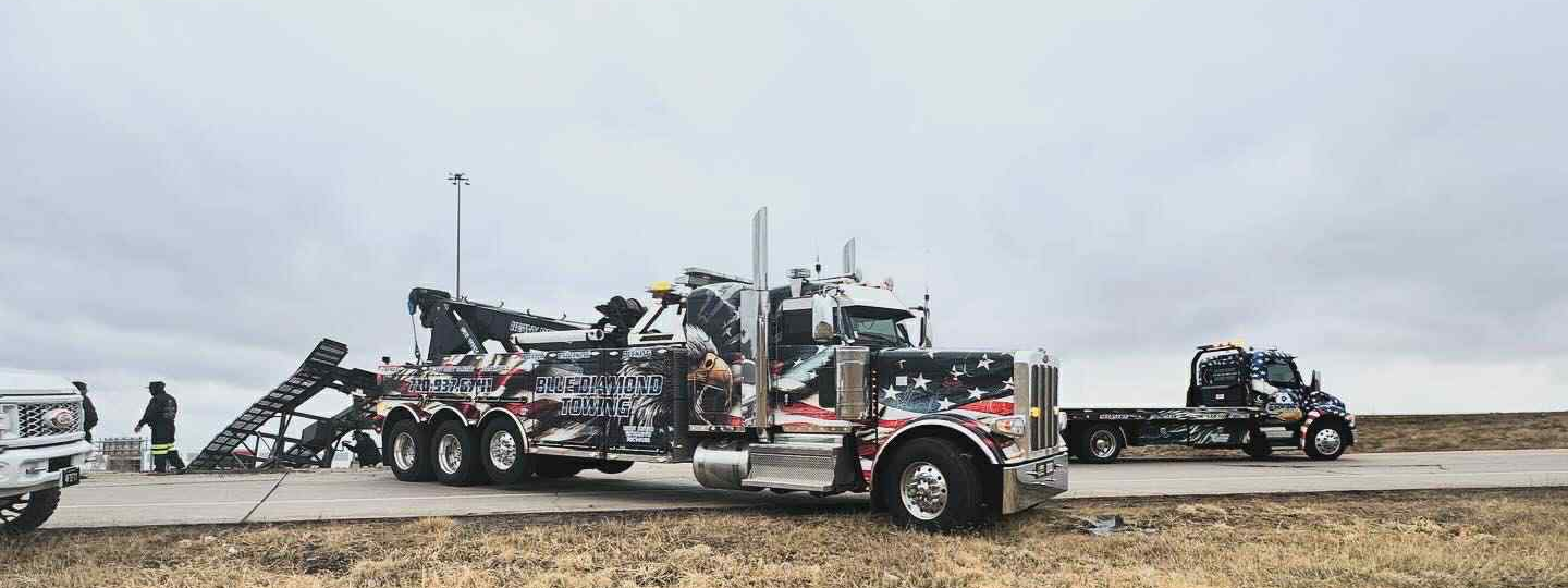 Tow trucks on a road under a cloudy sky; one has its arm extended, two people stand nearby.